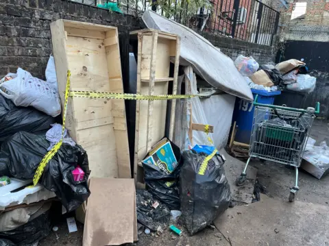 A pile of rubbish include over-spilling black bags are piled up next to wooden crates, other bins and a shopping trolley