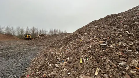 BBC A close up image of a large pile of waste which extends out into the distance of the photograph. The waste is made up of wood, plastic and other bits of household waste. There is a yellow JCB truck in the background. 