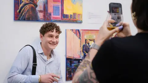 Irina Mackie Robbie Jefcott, now represented by Lux Gallery, stands next to paintings displayed on a gallery wall. He has a bag over his shoulder, a gold watch and a glass in his hand. Someone is standing in front of him taking his photograph with the artwork.