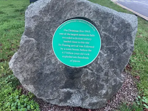 A large grey stone with a green circle plaque in the middle which reads "On Christmas Eve 1965, one of the largest meteorite falls recorded in British history landed close to this site. Its flaming arrival was followed by a sonic boom, before the 4.5 billion year old rock exploded into hundreds of pieces".