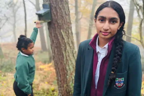 Schoolgirl sisters Avni and Vani are pictured in woodland, Vani looking at the camera, and Avni side-on with her hand up to a squirrel box on a tree.