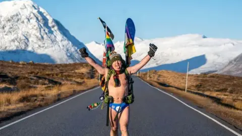 PA Media Mick Cullen, otherwise known as Speedo Mick, walks through Glencoe in sub-zero temperatures wearing only his swimming trunks. He is wearing his blue Speedos, a woolly hat, scarf, backpack, gloves and nothing else, his face tilted upwards to the sky with his eyes closed and a look of relief, against a background of snowy mountains in Glencoe, Scotland, in 2023