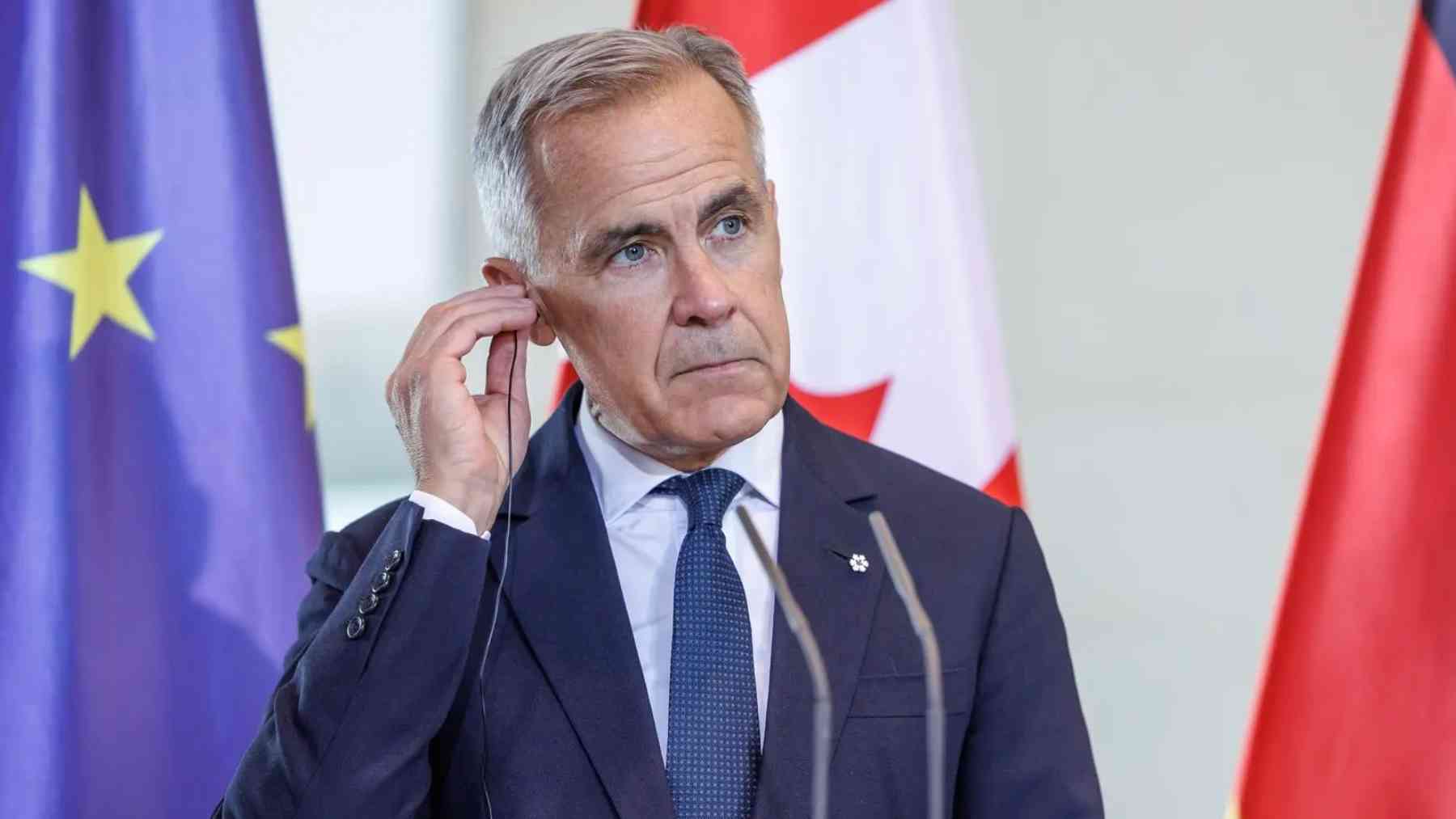 Government official at a podium adjusts an earpiece, with EU, Canada and Germany flags behind him.