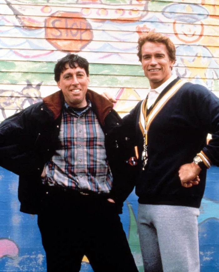 Two people standing in front of a colorful mural. One wears a plaid shirt and jacket, the other sports a sporty sweater and whistle