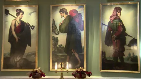 Three portraits hang on a wall in gold rimmed frames. The portraits show men in green and red clothing. 