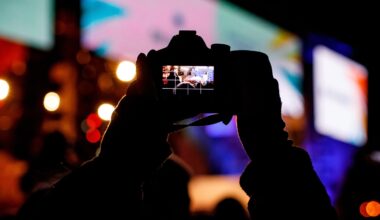 A person holds up a camera at night, capturing a brightly lit scene with colorful lights and blurred shapes in the background. The camera’s display screen shows the focused image being taken.