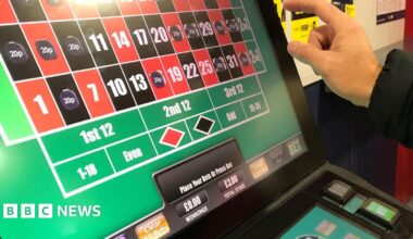 A man's hand is pictured about to press the brightly lit screen of a slot machine. The screen shows a series of red and black squares, some of which are filled with the price of bets placed. Details of stakes and winnings are also pictured.