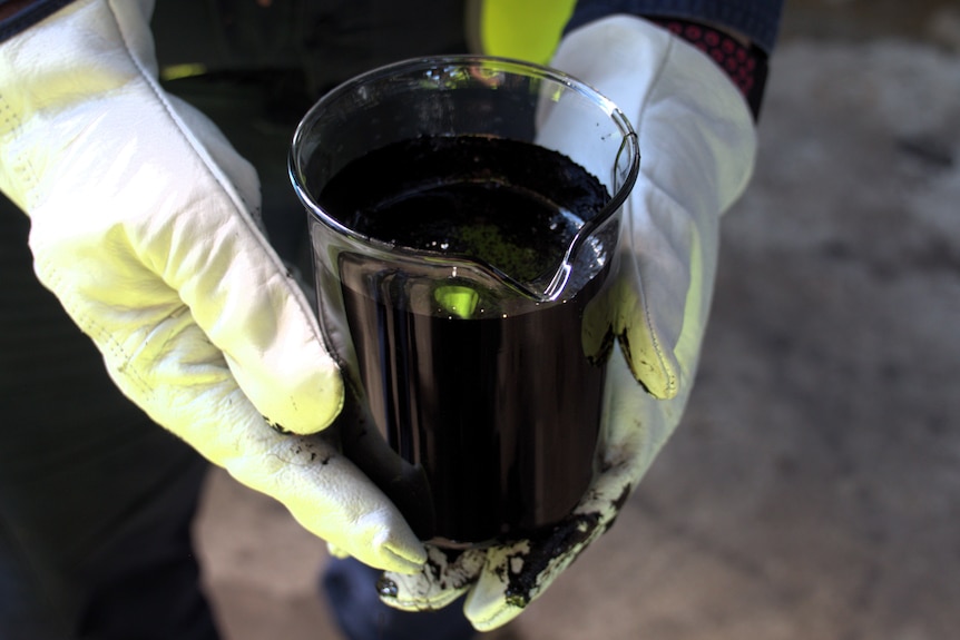 A glass beaker of black oil held by two hands in white gloves.