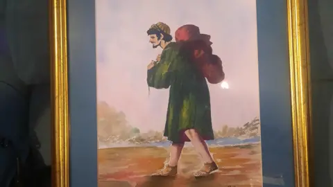 Oswald’s Hospice A watercolour of a man wearing a green coat, with a red bag on his back, and sandals, walking along a riverbank.