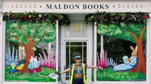 Maldon Books A shop front has a white sign at the top saying Maldon Books in a black font. The windows are full of paintings of trees, flowers and woodland creatures reading books. A woman wearing a high-vis top, tshirt, cap and sunglasses on her head stands with her arms up outside the shop.