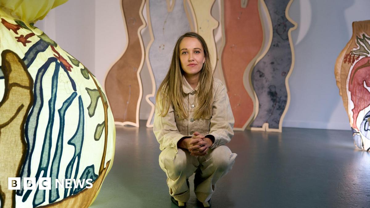 Bethany Williams wears a beige jacket and trousers, she has brown/ blonde hair and crouches in an exhibition which has colourful lights and arching coloured panels in the background.