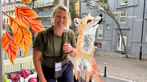 Art by Juliet Townsend A woman with blonde hair stands in a window and smiles at the camera. She is wearing a khaki tshirt and blue shorts. Behind her is a detailed painting of a fox and autumnal leaves in the window.