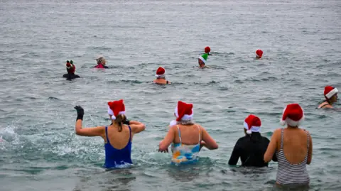 Getty Images The image shows a group of people swimming in open water. Several swimmers are wearing festive accessories such as red Santa hats and green elf hats. The swimmers appear to be close together, some with arms raised above the water, and most are wearing swimsuits or wetsuits. The water has some ripples.