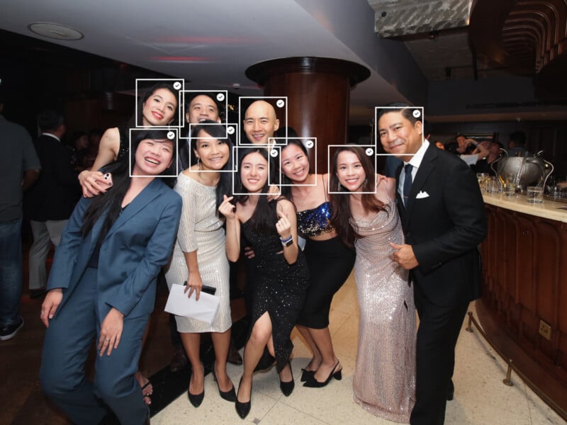 A group of eight people dressed in formal attire smile and pose together at an indoor event or party, with a curved wooden bar and dim lighting in the background.