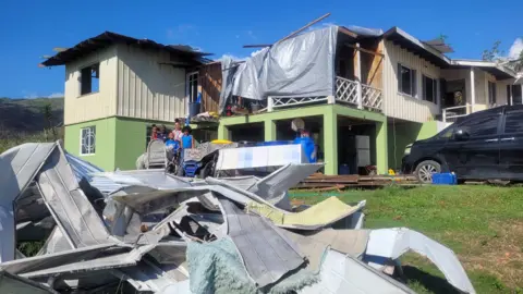 Handout A picture of the outside of the family home, which is green and cream. There is a grey tarpaulin covering a section of the roof. Metal scarps can been seen piled up in front of the home.