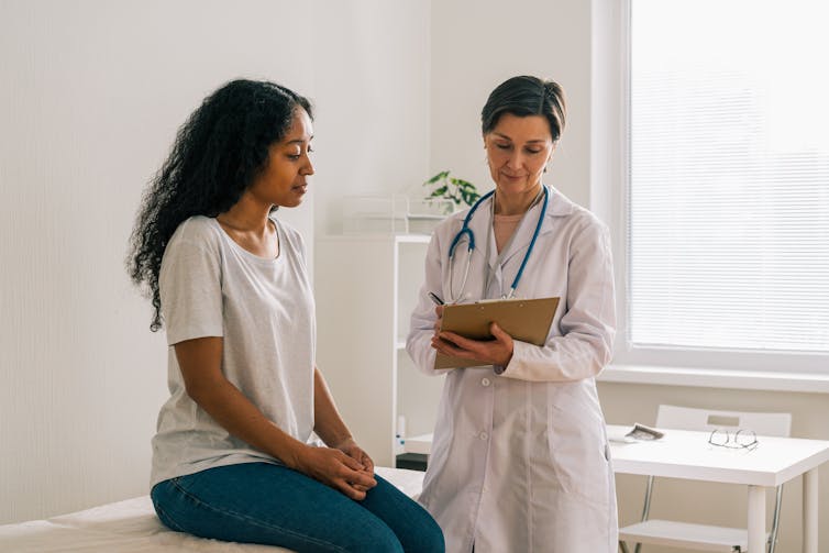patient sitting on table looks at doctor filling out form on clipboard