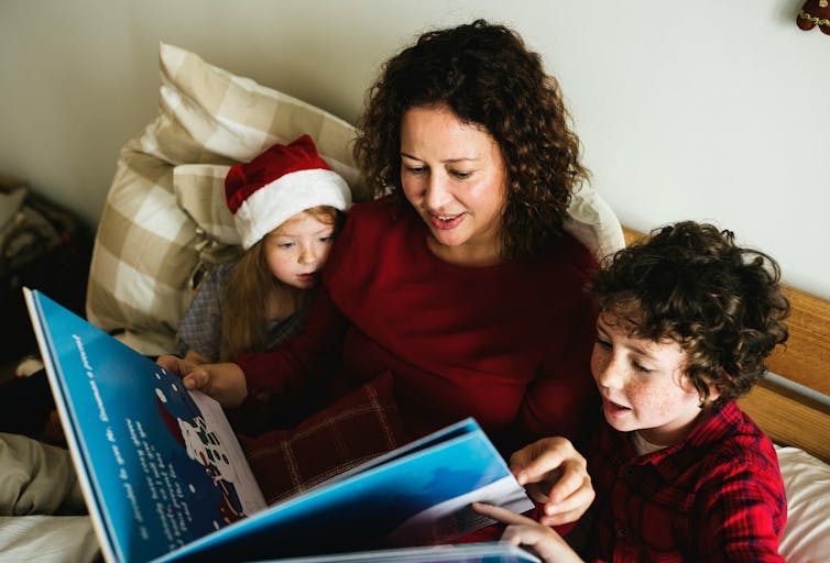 A woman and two children looking at a picture book. One of the children is wearing a Santa hat