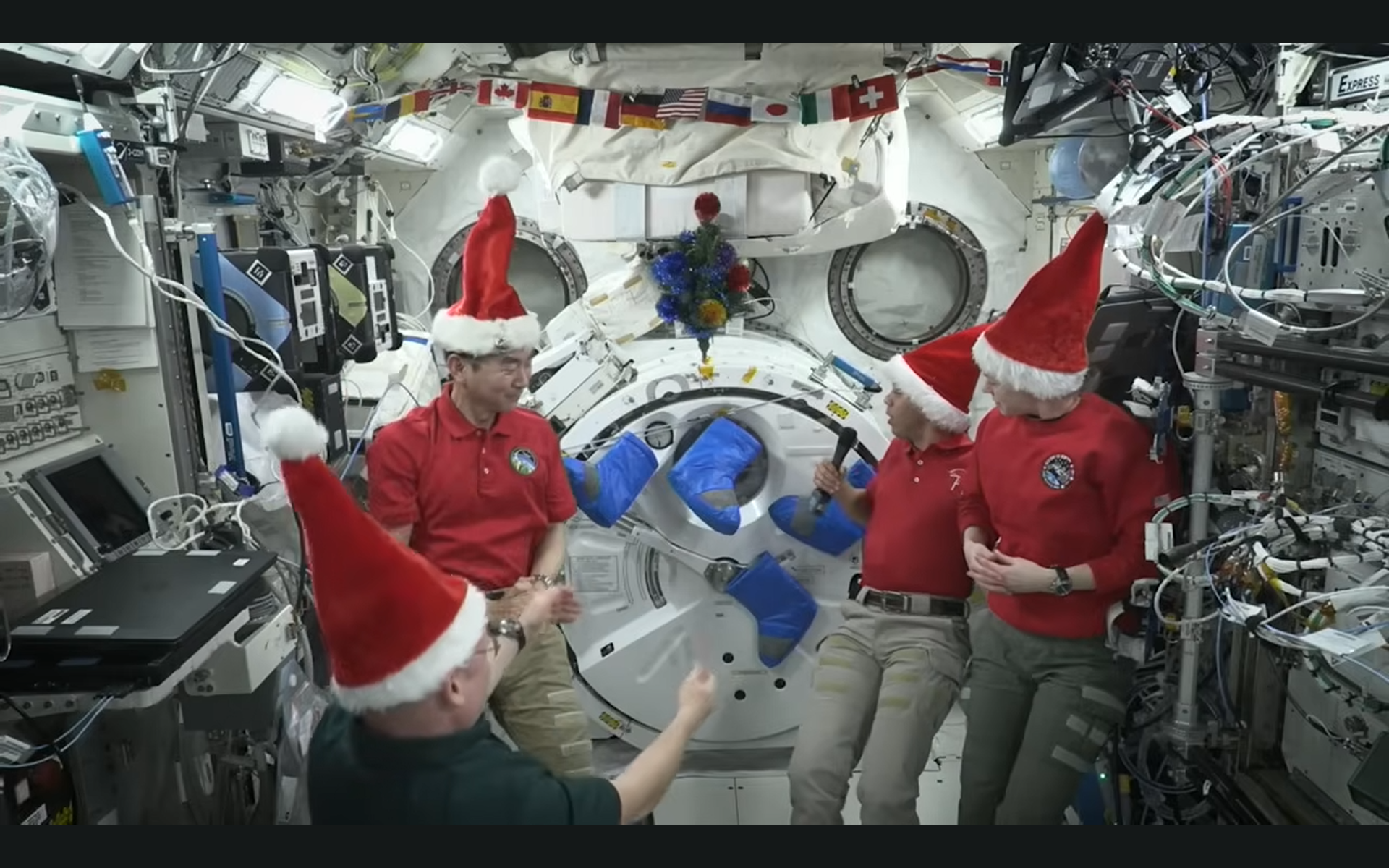 Four astronauts in Santa hats inside a space station decorated with space stockings and a Christmas tree.