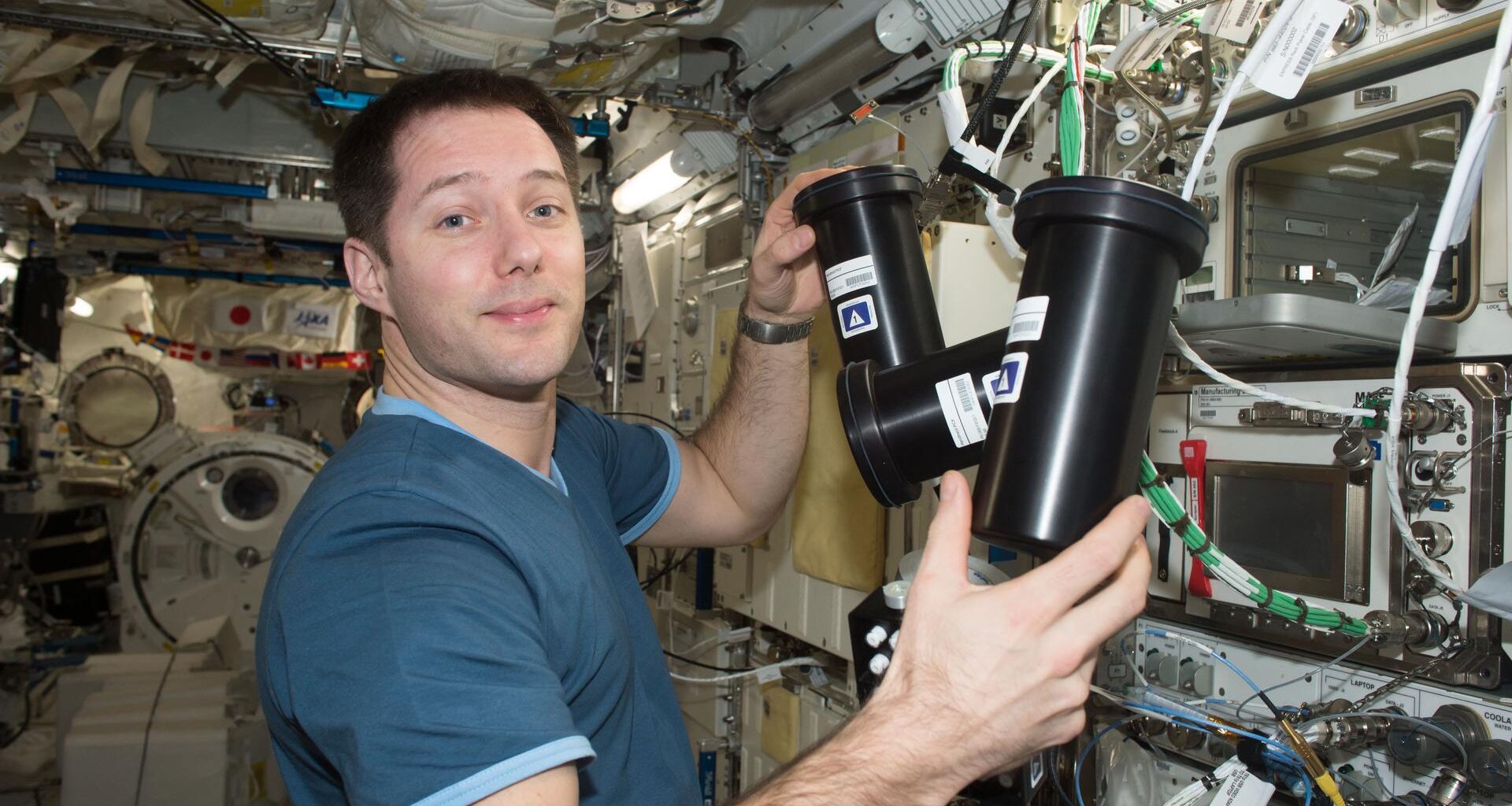 European Space Agency astronaut Thomas Pesquet works inside the International Space Station, . He is holding two cylindrical black experiment containers to deactivate and stow the Protein Crystal Growth-5 hardware. The background is filled withIn the background, cables, silver knobs, instruments, and research equipment are visible.