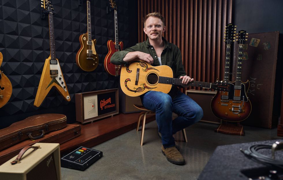 Musician sitting with an acoustic guitar in a room filled with various guitars and equipment.