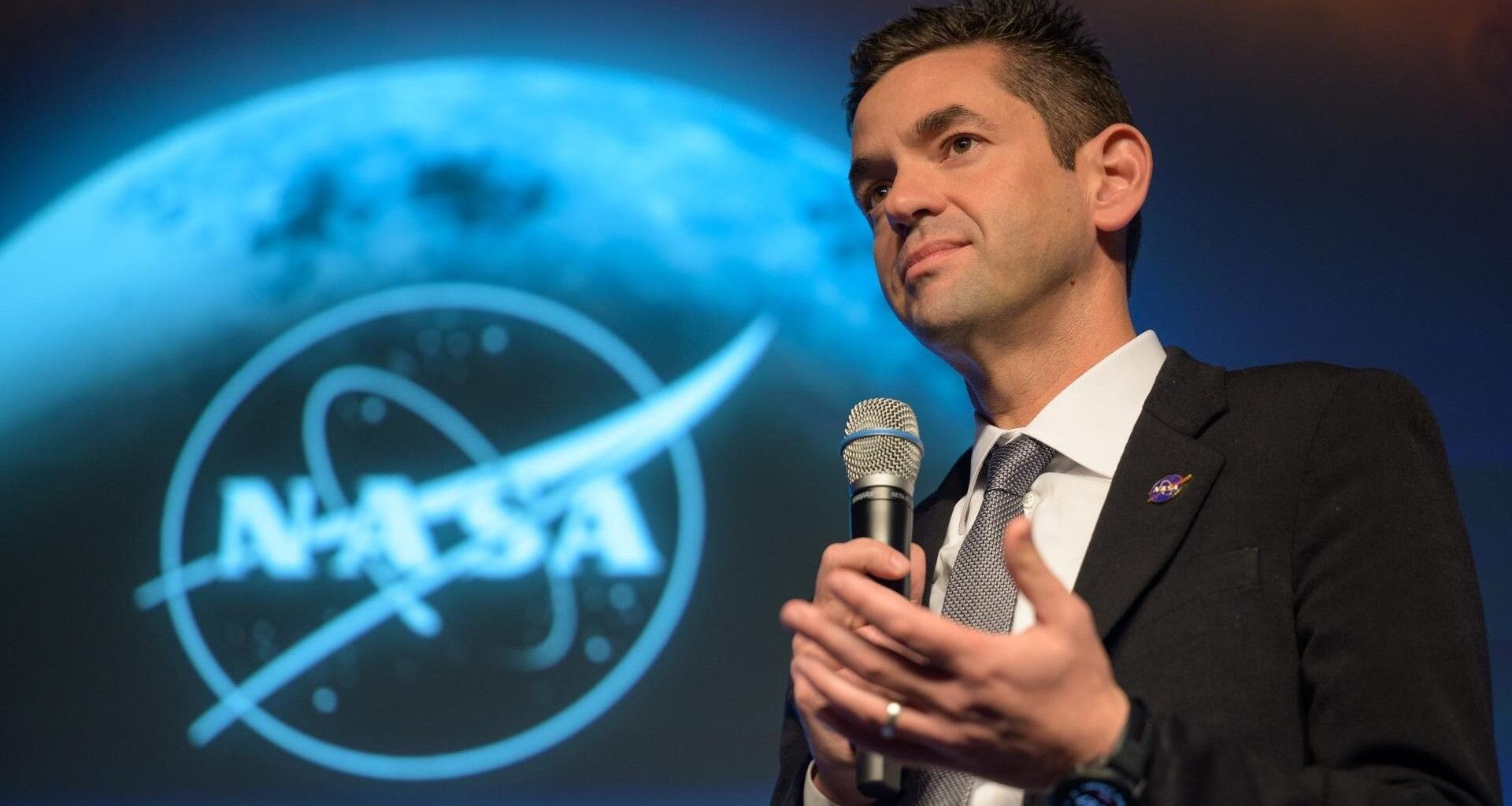 a man in a suit speaks into a microphone in front of a nasa emblem