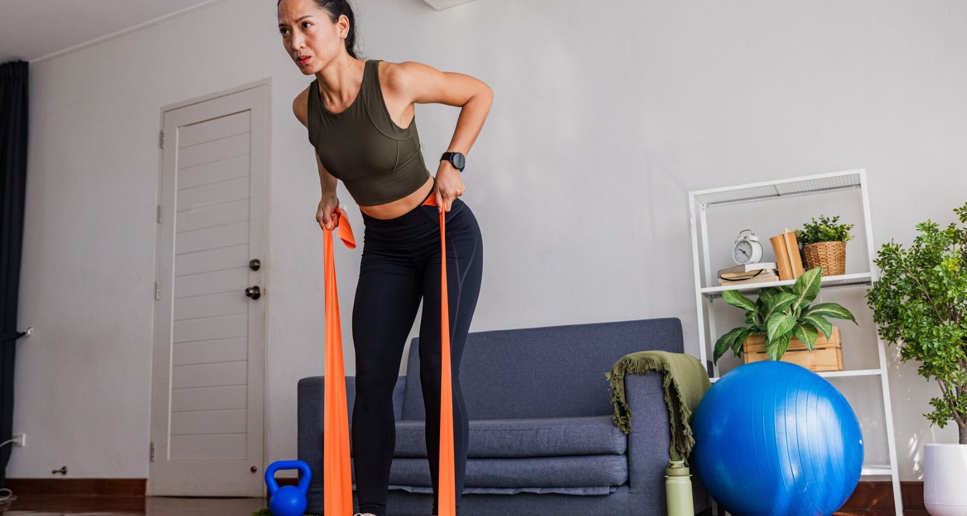 woman in a living room with an orange resistance band under her feet pulling the ends towards her in a bent-over row. there's a blue sofa and big blue balance ball next to her.