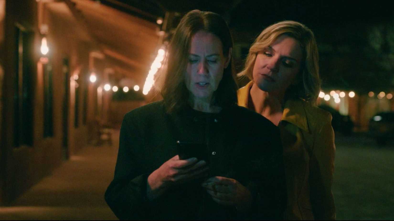 Miriam Shor as Helen and Rhea Seehorn as Carol Sturka. The two are in a car lot, lit up by lights. Carol looks over Helen's shoulder at the phone in Helen's hand.