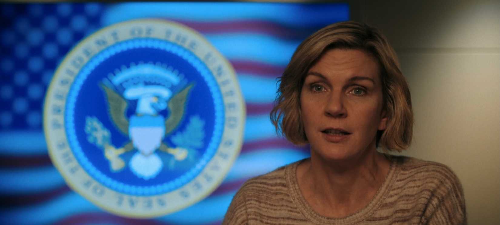 Carol (Rhea Seehorn) looks concerned while standing in front of the Presidential Seal of the United States in Pluribus