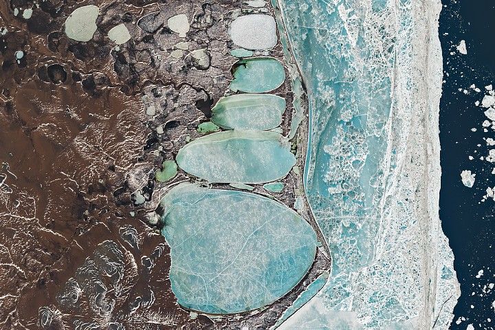 A Landsat image of Russia’s Chukchi Peninsula shows a narrow coastal sandspit separating the Arctic Ocean from a chain of elongated, ice-covered coastal lagoons near the village of Billings and Cape Billings. The pale blue lagoons vary in size and line up end-to-end, creating a shape that resembles a snowman pressed against the shoreline. Offshore, fractured sea ice forms a bright, textured band along the coast, while darker tundra and thermokarst lakes extend inland.