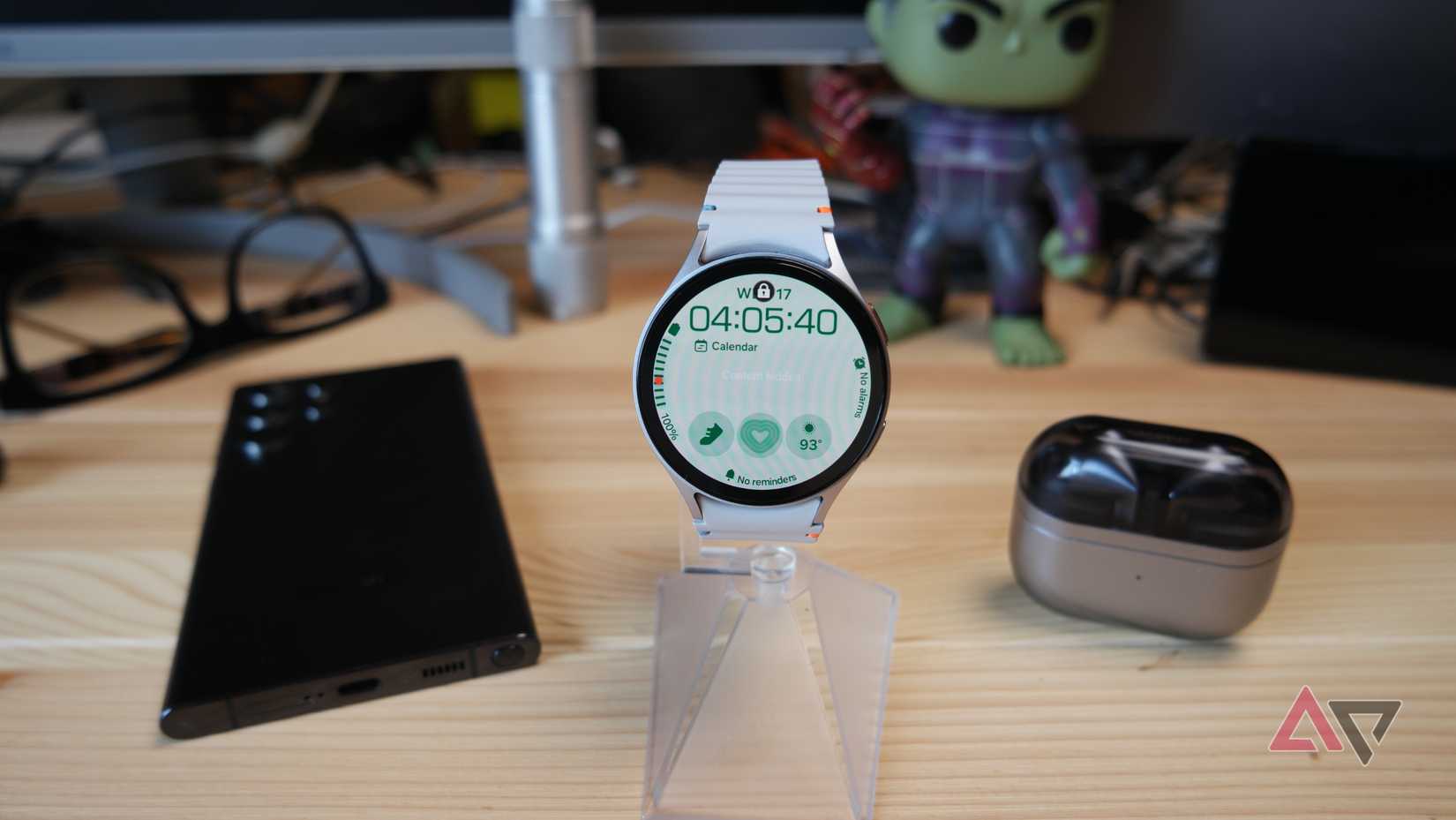 Samsung Galaxy Watch 7 on a stand, showing the home screen with other Galaxy products in the background