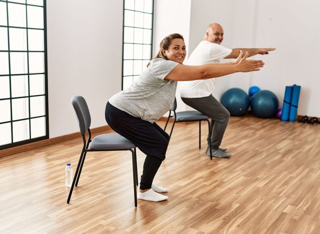 Middle age hispanic couple stretching using chair at sport center.
