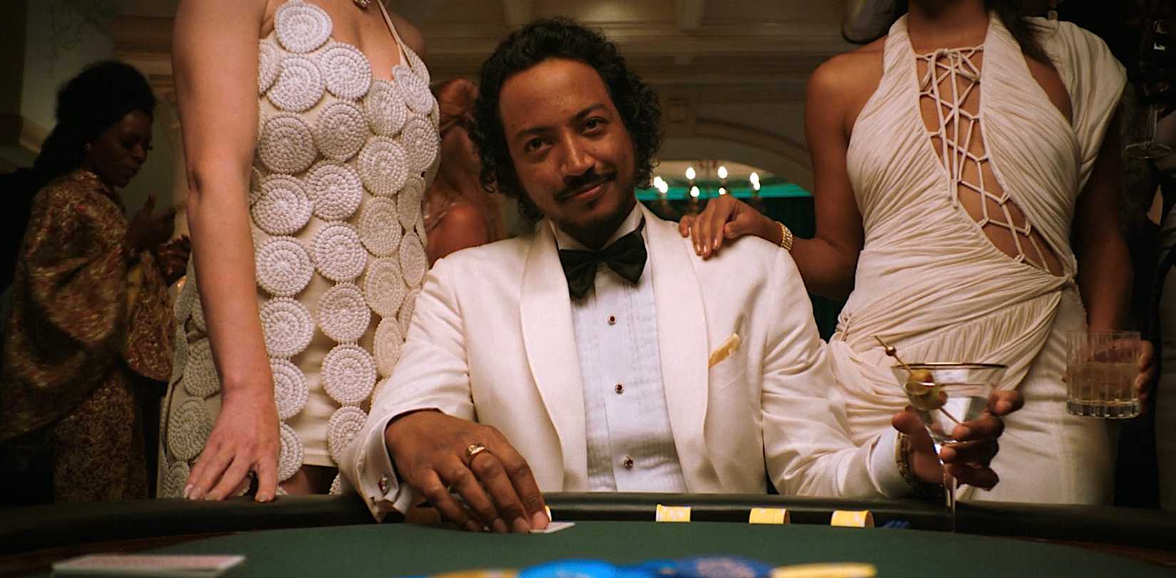 Koumba Diabaté (Samba Schutte), in a white tux and black bow tie, smirks as he sits at a Vegas poker table, holding a martini and flanked by beautiful women in episode 6 of Pluribus