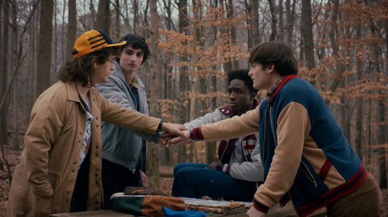 Dustin Henderson (Gaten Matarazzo), Mike Wheeler (Finn Wolfhard), Lucas Sinclair (Caleb McLaughlin), and Will Byers (Noah Schnapp) join hands in a forest in Stranger Things