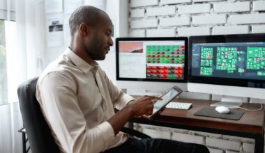 A person looking at a smartphone with computer screens in the background, showing stock prices.