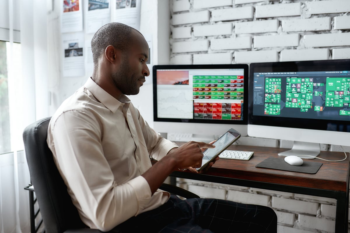 A person looking at a smartphone with computer screens in the background, showing stock prices.