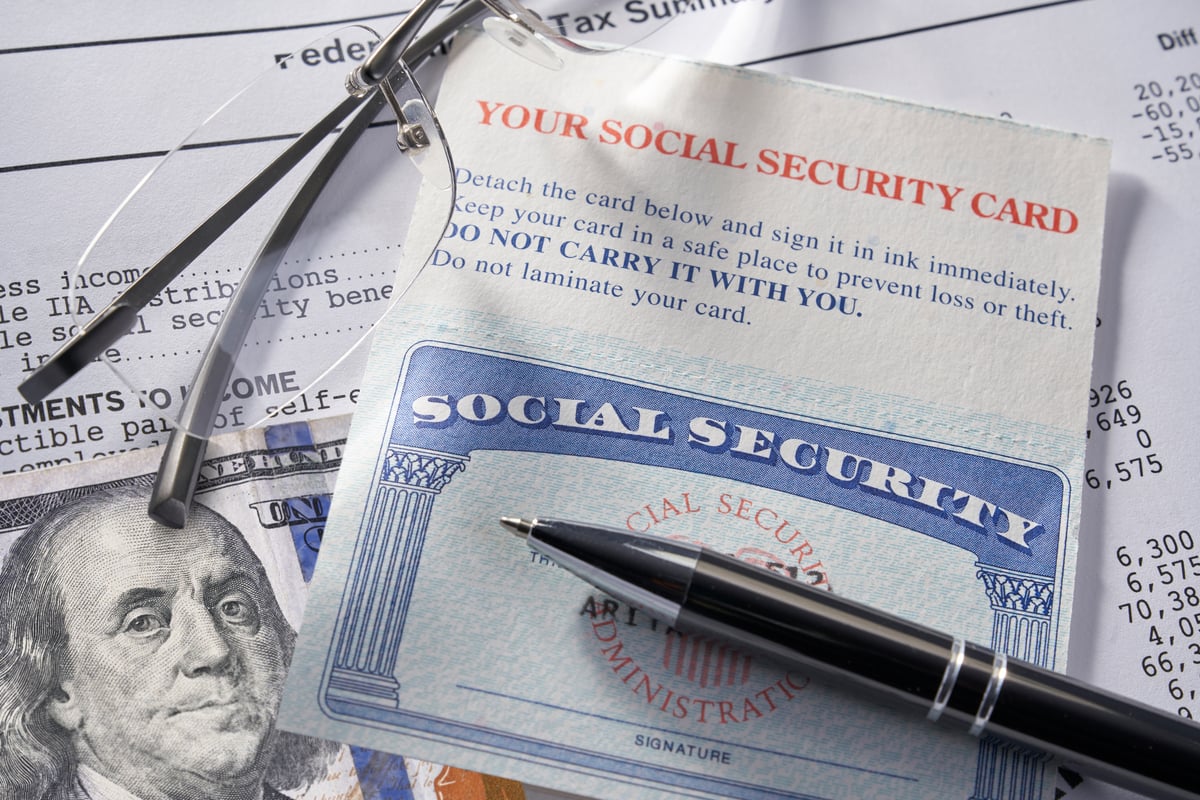A Social Security card laying on a financial statement with a pen, glasses, and $100 bill.