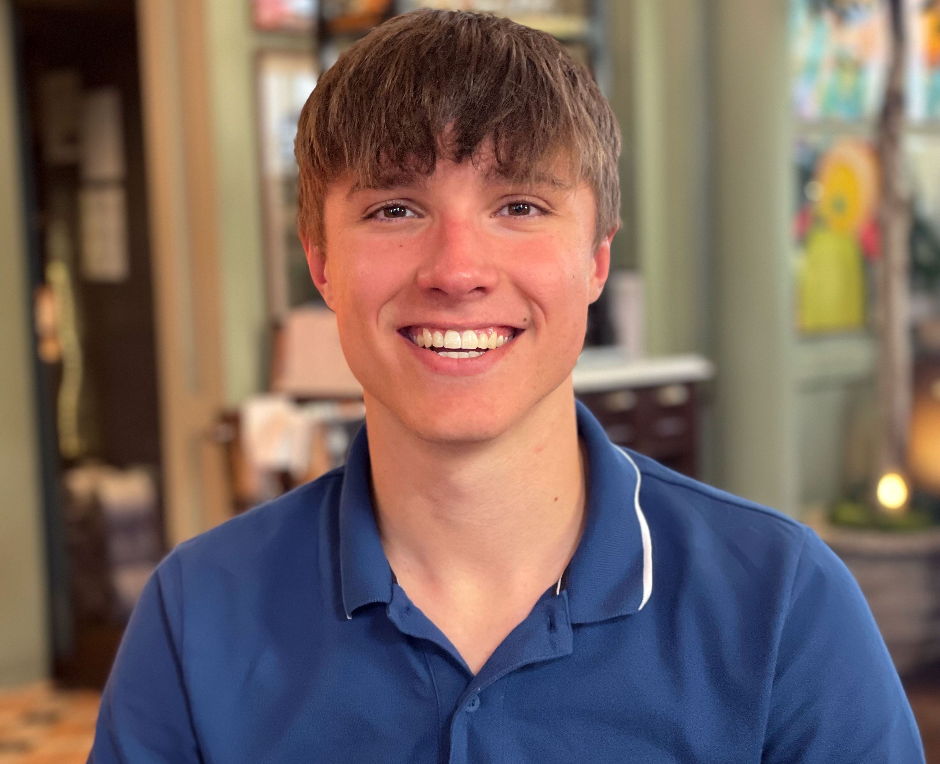 Barnaby Webber, a young man with short brown hair, smiles at the camera while wearing a navy blue polo shirt with "CK" embroidered on it.