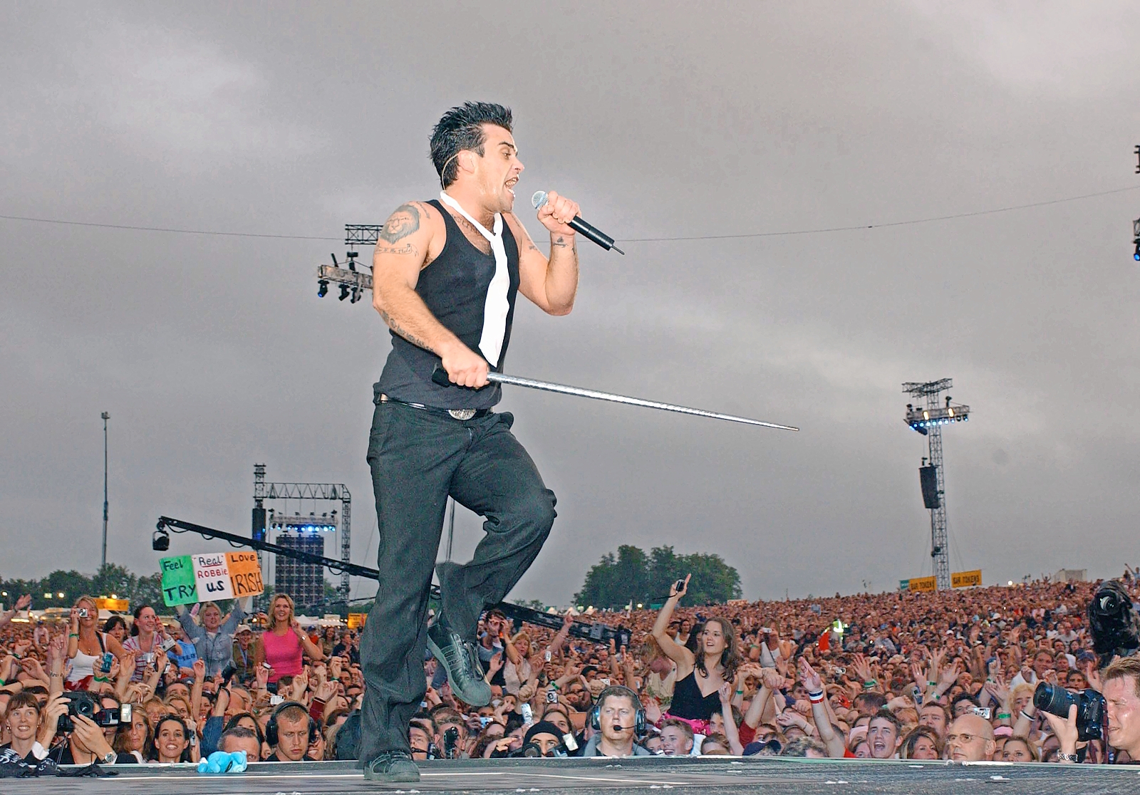 Robbie Williams performing with a microphone and a cane to a large crowd.