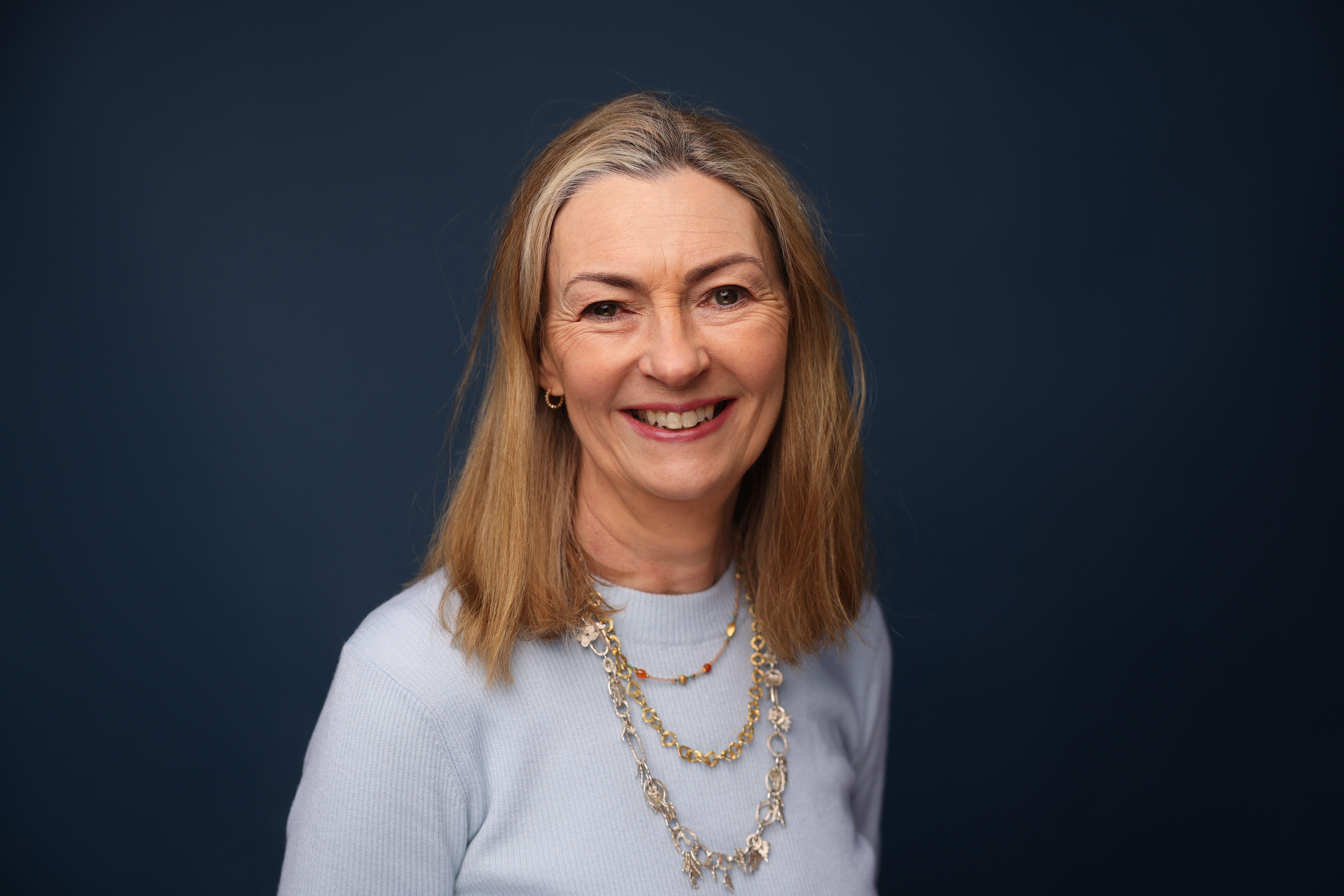 Anne Richards, CEO of Fidelity Intl Ltd., smiling in a light blue sweater with multiple necklaces, against a dark blue background.