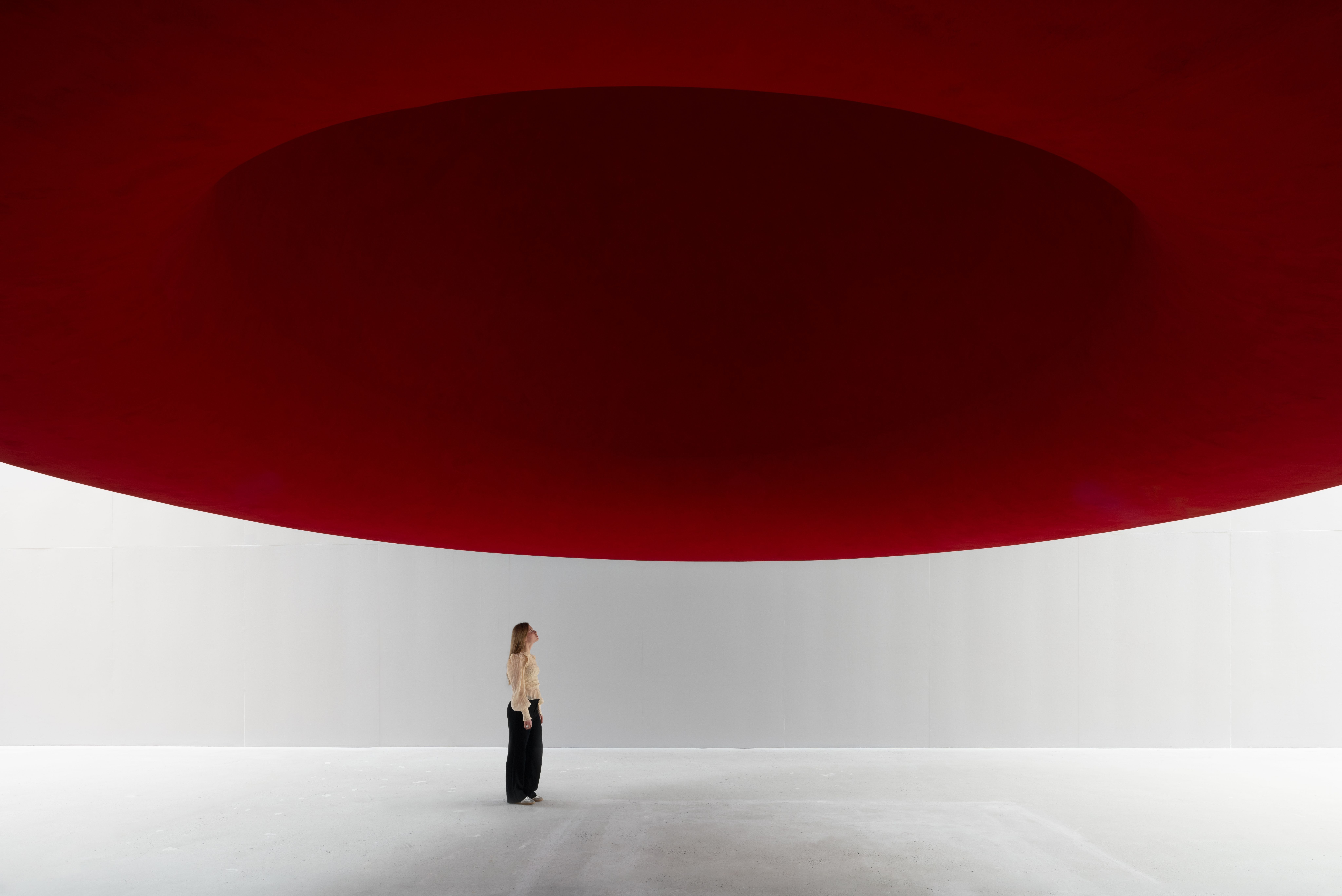 A woman stands in a large white room, looking up at a vast, dark red circular structure above her.