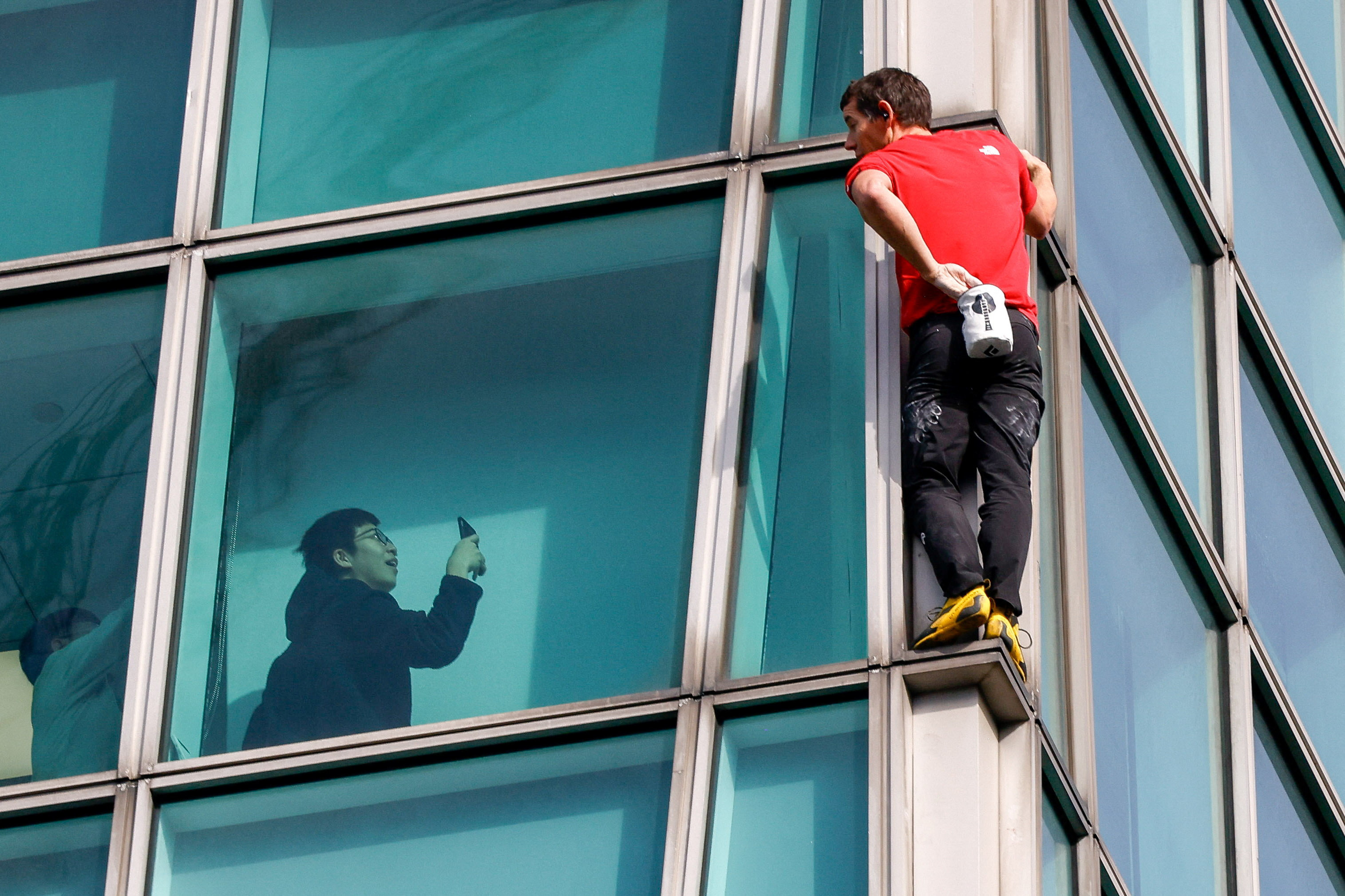 Climber Alex Honnold free soloing Taipei 101 Skyscraper with an onlooker taking a photo from inside.