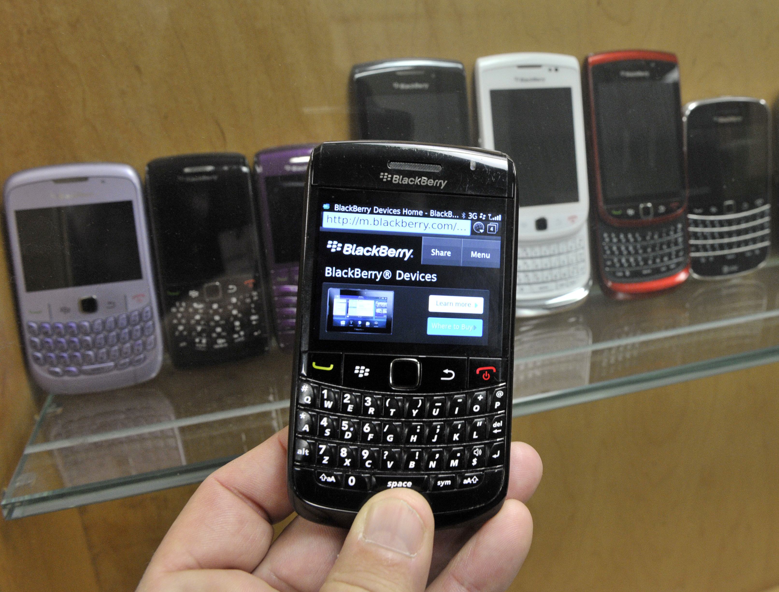 A person's hand holding a BlackBerry device displaying the "BlackBerry Devices Home" page, with several other BlackBerry devices in various colors visible in the background inside a glass cabinet.