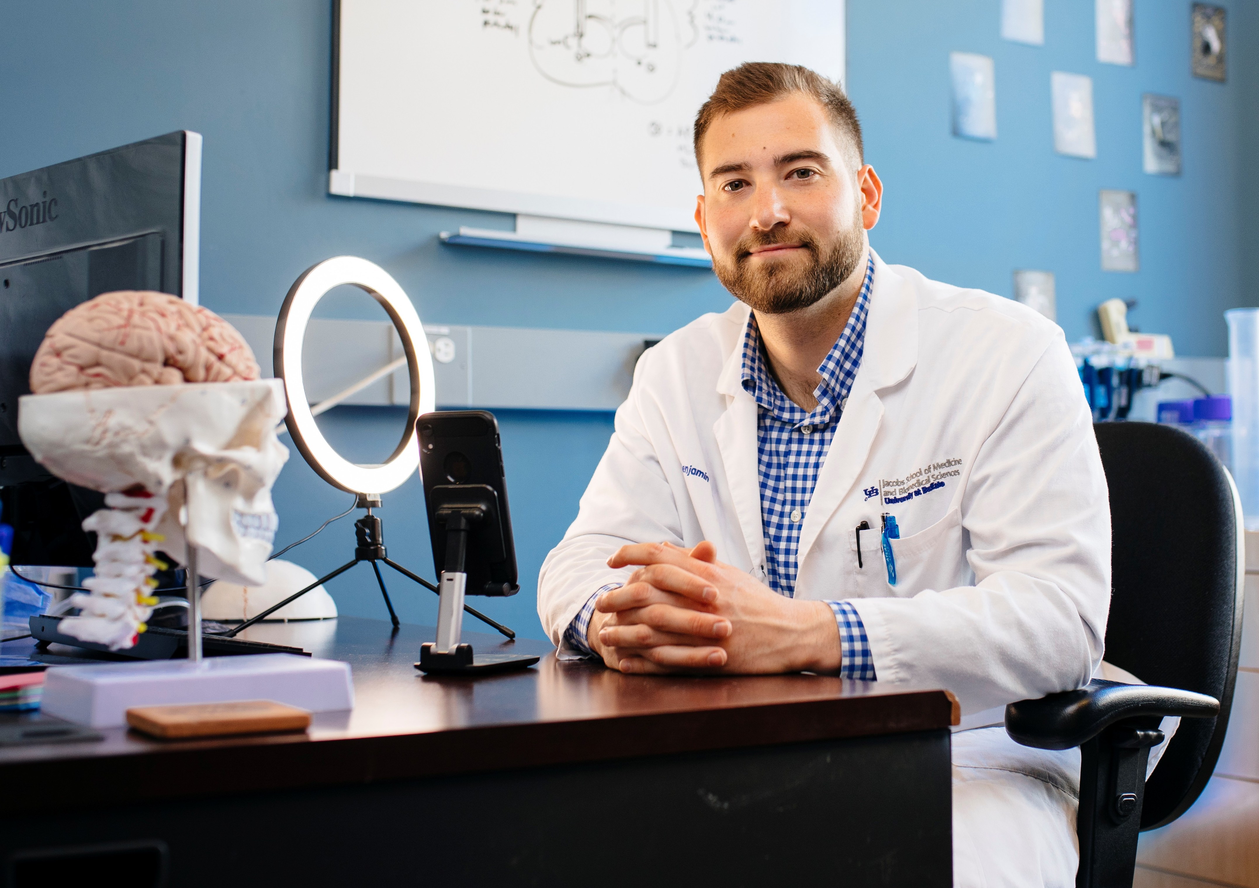 Dr. Ben Rein in a white lab coat, seated at his office desk with a brain and skull model, a ring light, and a whiteboard behind him.