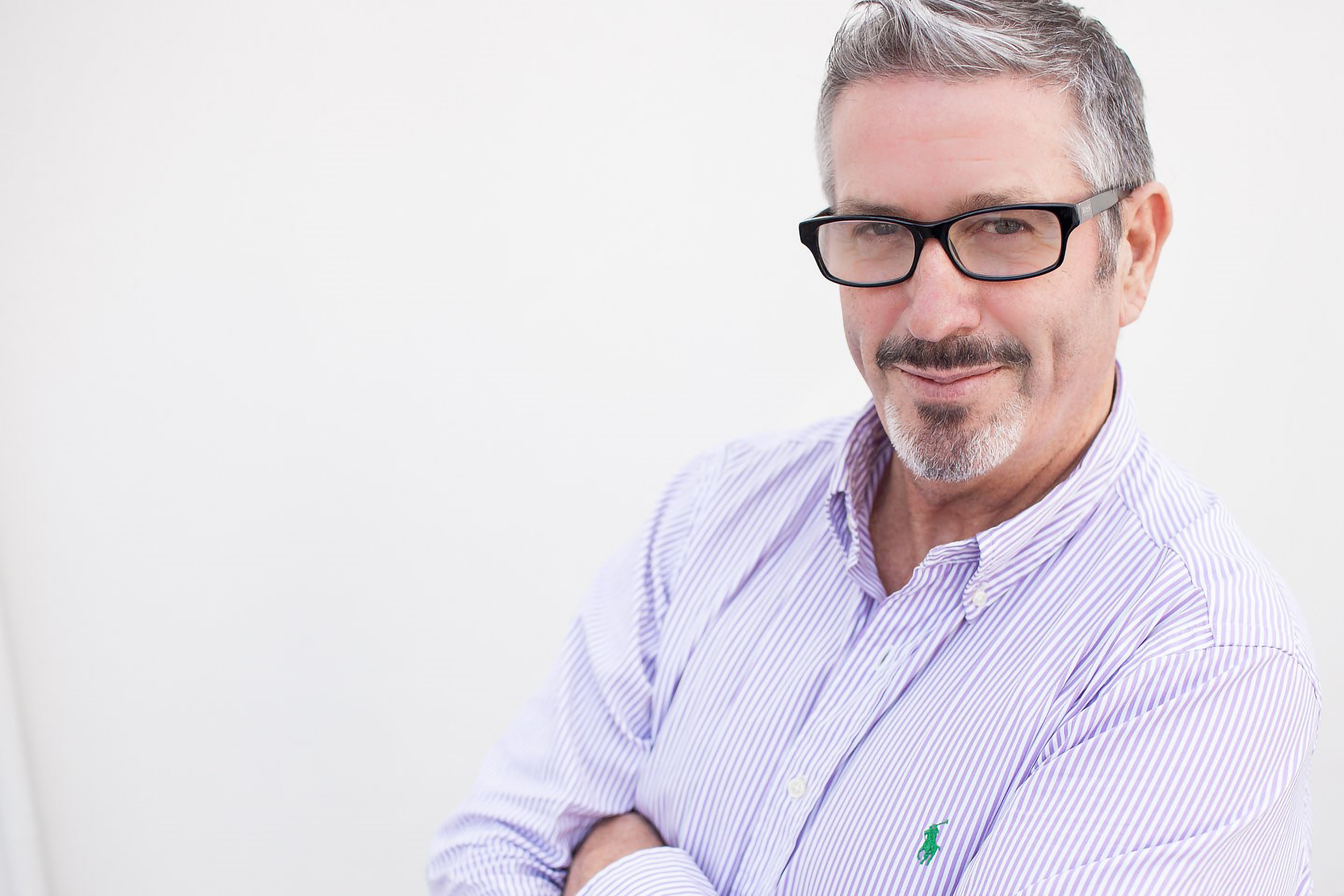 Garry Trainer, a man with graying hair and a beard, wearing glasses and a striped purple shirt, smiles at the camera.