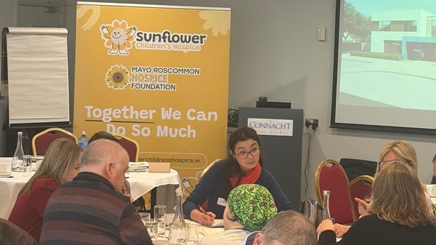 People sit at tables with a large sign reading Sunflower Children's Hospice in the background