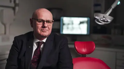 A bald man in glasses and a suit sits on a red dentist's chair in a surgery. There is a blurred computer screen in the background, showing what looks like an x-ray of teeth.