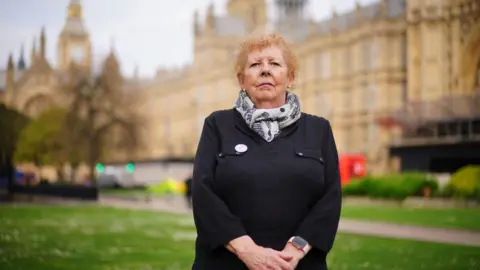 PA Media A woman wearing a black coat and patterned scarf standing in front of the Houses of Parliament.