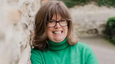 BAA Claire standing outdoors next to a stone wall. She is wearing a bright green turtleneck sweater and dark trousers. Her arms are crossed casually in front of her chest, and she is leaning slightly against the wall. The background features a gravel pathway and greenery, including ivy-covered stonework.