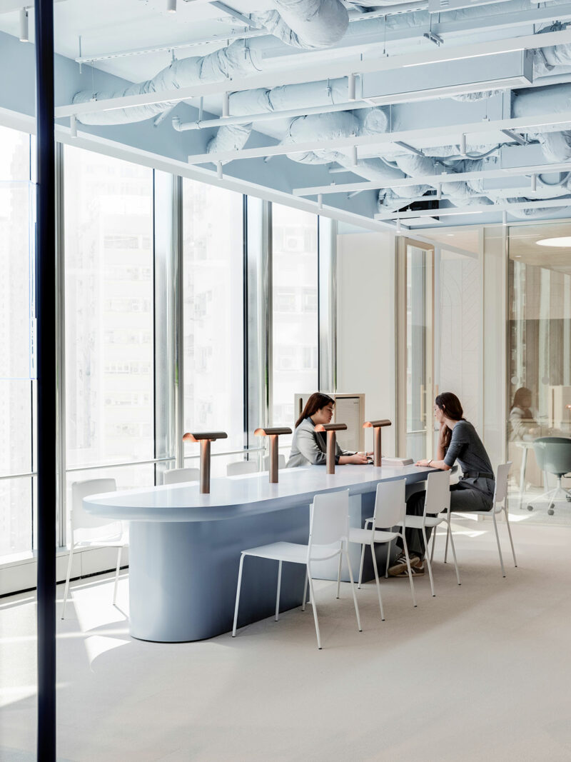 Two people sit across from each other at a long table in a modern, bright office space with large windows and exposed ceiling pipes.