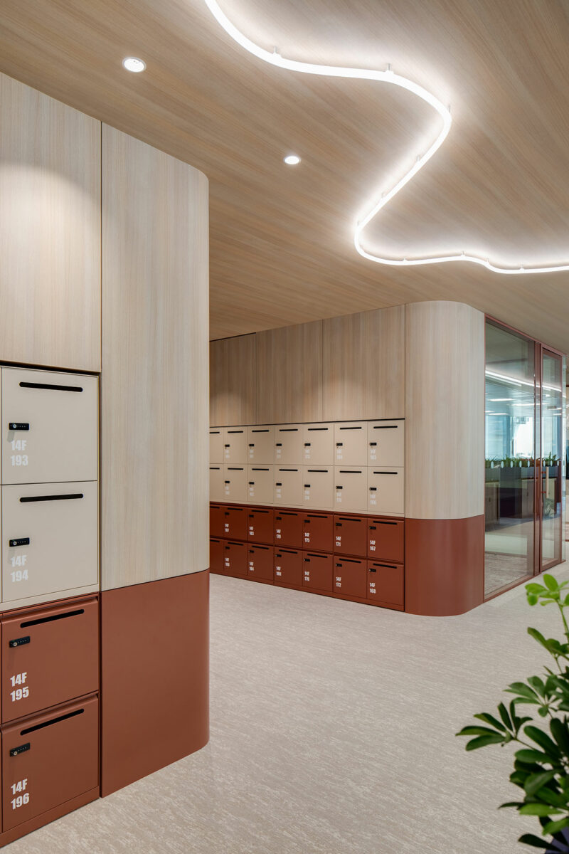 Modern office interior with beige and brown lockers, a curved light fixture on the ceiling, and glass-walled meeting room in the background.